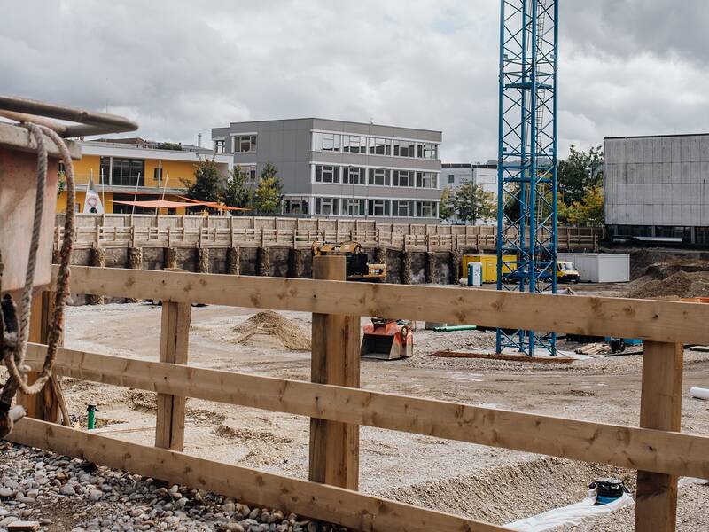 Eine Baustelle mit einem Baukran, einem Schüttbehälter und umliegenden Gebäuden im Hintergrund.